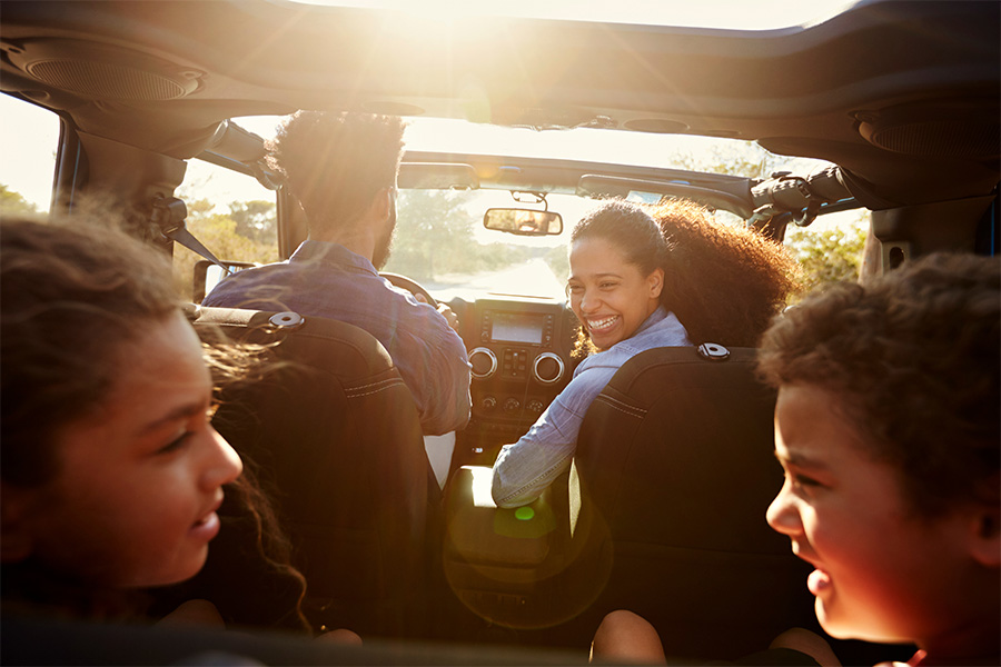 Car Insurance - Father Driving a Car with Open Windows as Mother Looks Back Smiling at Her Kids in the Back Seat on a Beautiful Sunny Day